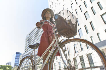 Nachdenkliche Frau steht mit Fahrrad gegen Gebäude und Himmel an einem sonnigen Tag - VEGF02491