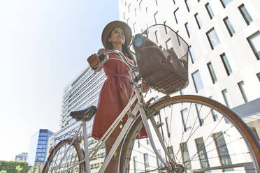 Thoughtful woman standing with bicycle against buildings and sky during sunny day - VEGF02491
