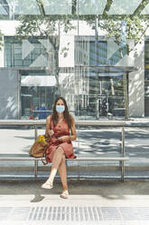Woman wearing mask while sitting at bus stop in city - VEGF02494