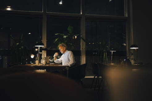 Nachdenklicher Geschäftsmann, der bis spät in die Nacht arbeitet, während er mit seinem Laptop an einem dunklen Arbeitsplatz sitzt - MASF19370