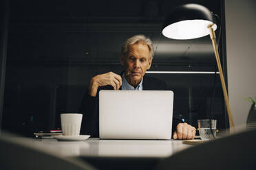 Engagierter älterer männlicher Unternehmer, der auf seinen Laptop schaut, während er spät in einem kreativen Arbeitsplatz arbeitet - MASF19381