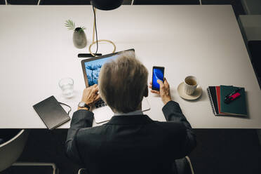 Hohe Winkel Rückansicht der älteren Geschäftsmann mit Smartphone und Laptop während der Arbeit spät am Schreibtisch im Büro - MASF19382