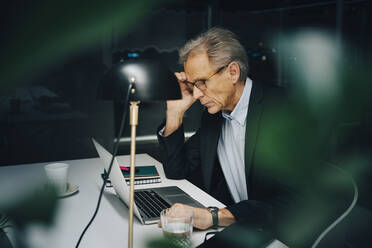 Besorgter älterer Geschäftsmann arbeitet bis spät in die Nacht, während er mit seinem Laptop im Coworking Space sitzt - MASF19385