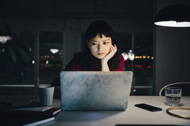 Engagierte junge Geschäftsfrau mit Hand am Kinn, die einen Laptop benutzt, während sie spät im Büro arbeitet - MASF19388