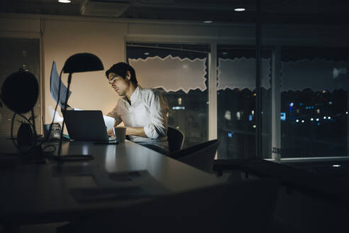 Engagierter männlicher Unternehmer, der ein Dokument liest, während er spät in einem dunklen Coworking Space arbeitet - MASF19396