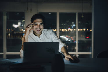 Lächelnder männlicher Berufstätiger, der noch spät arbeitet, während er auf seinen Laptop in einem dunklen Coworking Space schaut - MASF19397