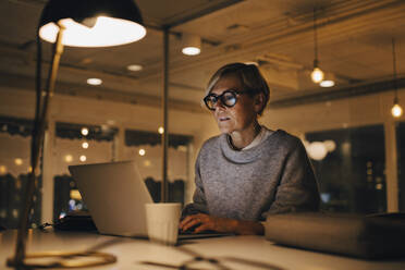 Confident mature female business professional using laptop while sitting at desk working late in office - MASF19422