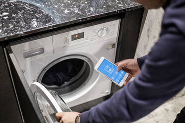 Ausgeschnittenes Bild eines Mannes, der eine Waschmaschine mit einer mobilen App im Smart Home steuert - MASF19423