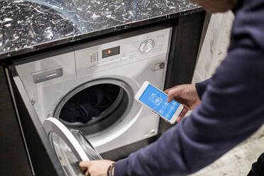 Cropped image of man controlling washing machine with mobile app at smart home - MASF19423