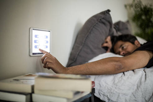 Mann benutzt ein an der Wand befestigtes digitales Tablet, während er auf dem Bett im Smart Home liegt - MASF19432