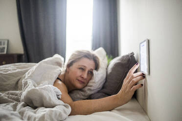 Frau benutzt ein an der Wand befestigtes digitales Tablet, während sie auf dem Bett im Smart Home liegt - MASF19433