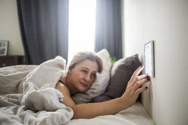 Woman using digital tablet mounted on wall while lying on bed at smart home - MASF19433