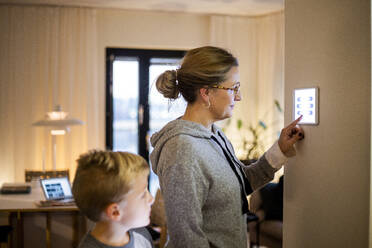 Sohn sieht seine Mutter an, die ein digitales Tablet an der Wand im Smart Home benutzt - MASF19453