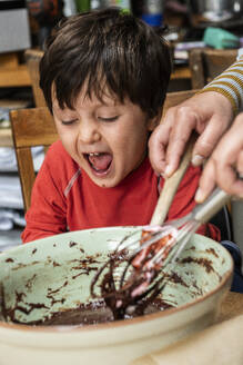 Boy with black hair sitting at a kitchen table, baking chocolate cake. - CUF55686