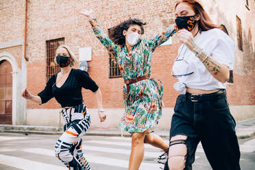 Three young women wearing face masks during Corona virus, running across a pedestrian crossing in a street. - CUF55728