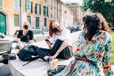 Three young women wearing face masks during Corona virus, sitting on a riverbank, chatting. - CUF55729