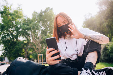 Junge Frau mit Gesichtsmaske während des Corona-Virus, sitzt im Freien und überprüft ihr Handy. - CUF55730