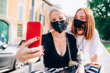 Two young women wearing face masks during Corona virus, sitting on a riverbank, taking selfie. - CUF55731