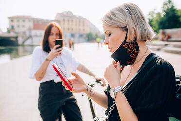 Zwei junge Frauen, die während des Corona-Virus Gesichtsmasken tragen, stehen im Freien und benutzen ein Mobiltelefon. - CUF55733
