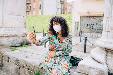 Junge Frau mit Gesichtsmaske während des Corona-Virus, sitzt im Freien und macht ein Selfie. - CUF55750