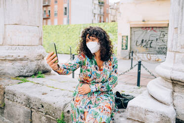 Young woman wearing face mask during Corona virus, sitting outdoors, taking selfie. - CUF55750