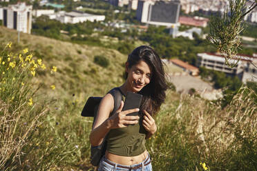 Young woman with long brown hair standing on hill above city, taking selfie with mobile phone. - CUF55755