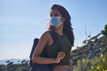 Junge Frau mit langen braunen Haaren steht auf einem Hügel, trägt einen Rucksack und einen Mundschutz während der Coronavirus-Krise. - CUF55756