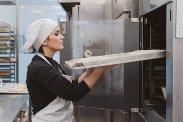 Bäckerin schiebt Backblech in den Ofen in einer Bäckerei - EBBF00370