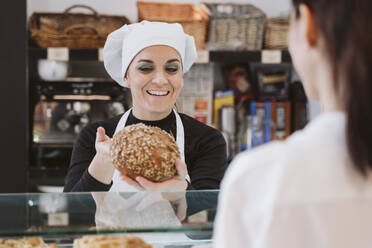 Glückliche Bäckerin, die einem Kunden in einer Bäckerei Vollkornbrötchen verkauft - EBBF00400