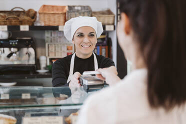 Happy female baker receiving contactless payment through credit card from customer at bakery - EBBF00403