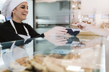 Happy baker receiving contactless payment through credit card from customer at bakery - EBBF00405