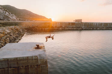 Side view of unrecognizable person doing back flip while jumping in sea water from concrete plate during magnificent sunrise on beach - ADSF00214