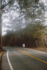 Rückansicht eines anonymen jungen Mannes, der auf einem Skateboard auf einer asphaltierten Straße in einem wunderschönen Wald an einem nebligen Tag in Big Sur, Kalifornien, fährt - ADSF00216