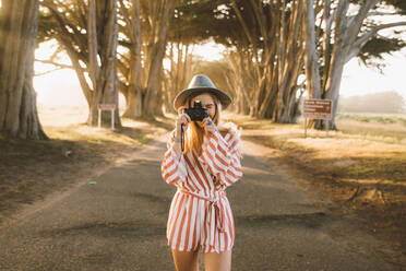 Junge Frau in stilvollem Outfit mit Fotokamera, um Bilder zu machen, während auf der Straße in erstaunlichen Baum-Tunnel am sonnigen Tag in der wunderschönen Natur stehen - ADSF00223