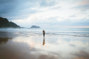 Back view of young woman standing on wet sand of beach near waving sea on cloudy day in Zarautz, Spain - ADSF00226