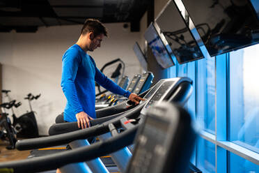 Side view of young man in blue sport pullover doing exercise on treadmill in gym - ADSF00235