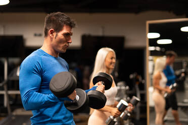 Side view of handsome man in blue pullover and attractive woman in sportswear standing in gym in front of mirror and doing exercise with dumbbells - ADSF00261