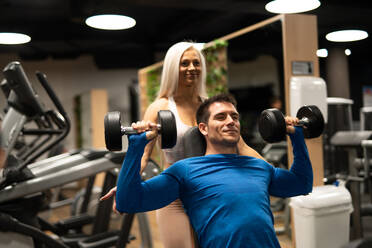 Schöne Frau mit blondem Haar, die einem Mann im blauen Pullover bei einer Übung mit Hanteln im Fitnessstudio hilft - ADSF00262