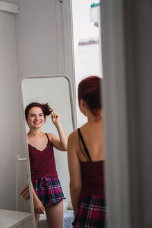 Rückenansicht einer attraktiven Frau im Schlafanzug, die in der Nähe eines Spiegels steht und ihr Haar im Zimmer berührt - ADSF00282