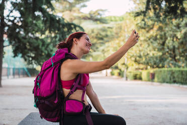 Seitenansicht einer attraktiven Frau in Sportkleidung mit rosa Rucksack, die im Park sitzt und ein Selfie mit ihrem Smartphone macht - ADSF00287