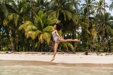 Side view cheerful attractive Hispanic woman with curly hairs jumping with legs to sides on shore with green forest in Bocas del Toro Islands, Panama - ADSF00298