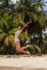 Young lady jumping on sand beach with tropical forest near water - ADSF00299