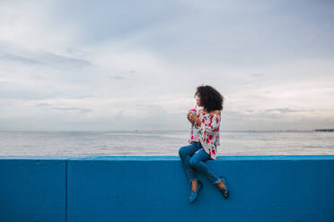 Ethnische stilvolle Frau, die ein köstliches Getränk in einer Tasse genießt, sitzend am blauen Zaun der Strandpromenade, Panama-Stadt - ADSF00304