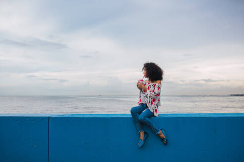 Ethnic stylish woman enjoying delicious drink in cup sitting on blue fence of seafront, Panama city - ADSF00304