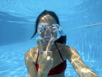 Mid adult woman gesturing peace sign while taking selfie in swimming pool - EGAF00426