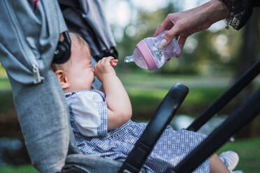 Hand of anonymous mother holding bottle of water and trying to comfort crying baby in stroller on blurred background of park - ADSF00308