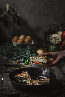 Blick auf die Hand mit leckeren Tortilla auf Pfanne neben Platten mit Scheiben, Tomaten, Früchte, Nüsse und Blätter auf Holzbrett - ADSF00328