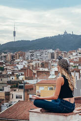 Seitenansicht einer attraktiven blonden Frau mit Kopftuch, die auf einem Dach in Meditationsposition sitzt und auf eine malerische Stadtlandschaft blickt - ADSF00411