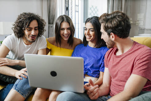 Lächelnde multiethnische Freunde teilen sich einen Laptop, während sie im Wohnzimmer sitzen - JSMF01593