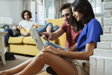Young couple sharing laptop while friends relaxing on sofa in living room at home - JSMF01595